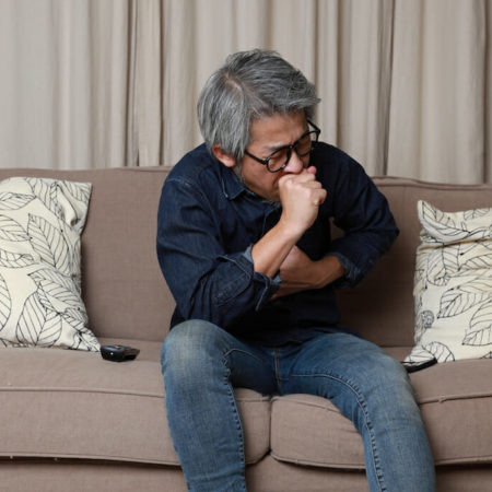 An older man sitting on couch while coughing from post nasal drip.