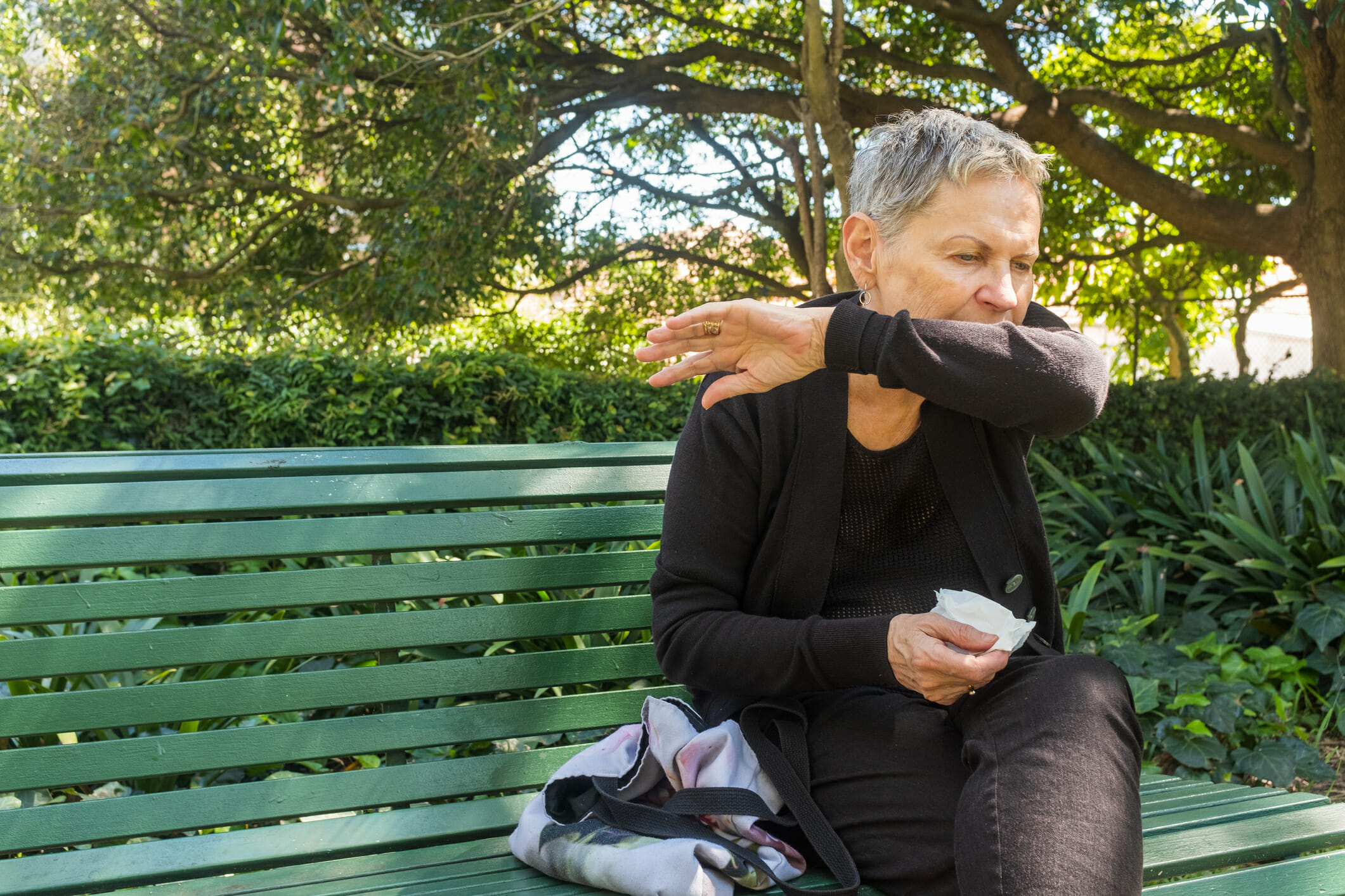 Elderly woman sitting on park bench outdoors coughing into elbow and holding tissue