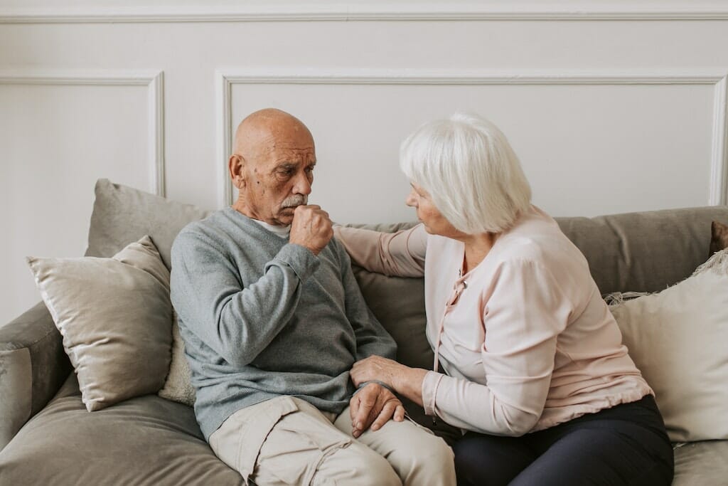 Older man coughing from bronchiectasis while sitting on couch with wife.